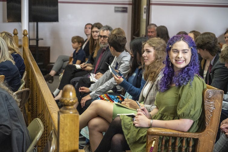 Young people fill a bench in a courthouse, some are elementary school age others in their teens. A girl at the end with purple hair smiles at someone across the aisle.