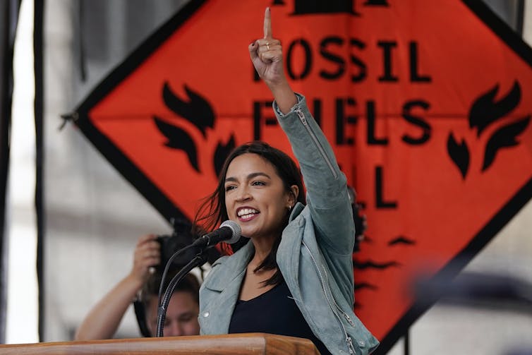 A woman speaking at a microphone points upward. Behind her is a sign reading: 'Fossil Fuels Kill'.