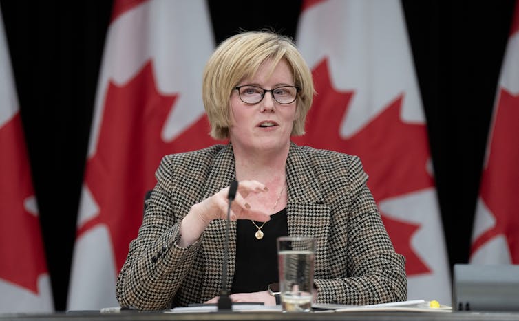 A middle-aged woman with short, blonde hair speaks into a microphone while seated in front of a row of Canadian flags