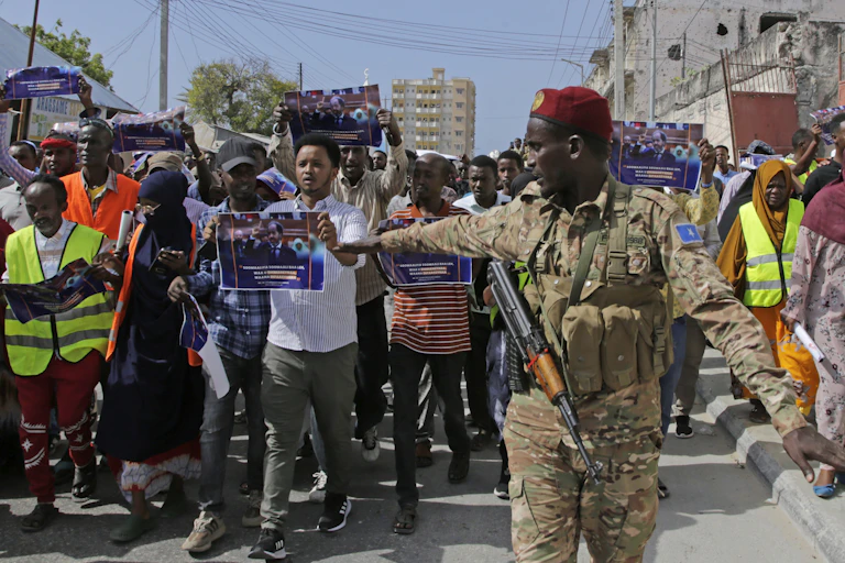 A man in army fatigues with a gun slung around his neck stands in front of a crowd of protestors.