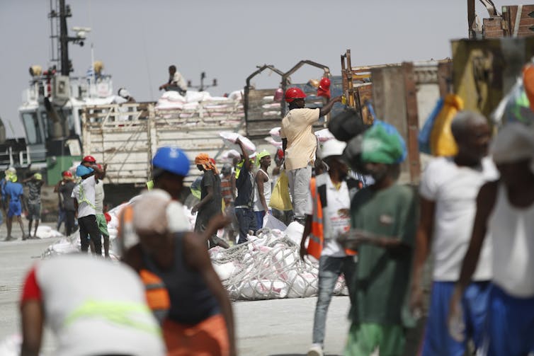 People walk around in bright colored clothing in front of a seaport.