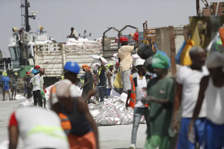 People walk around in bright colored clothing in front of a seaport.