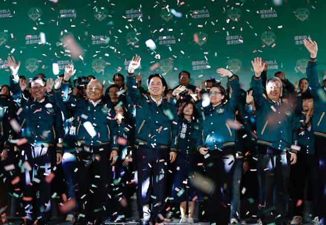 A large group of people in green jackets wave under falling confetti