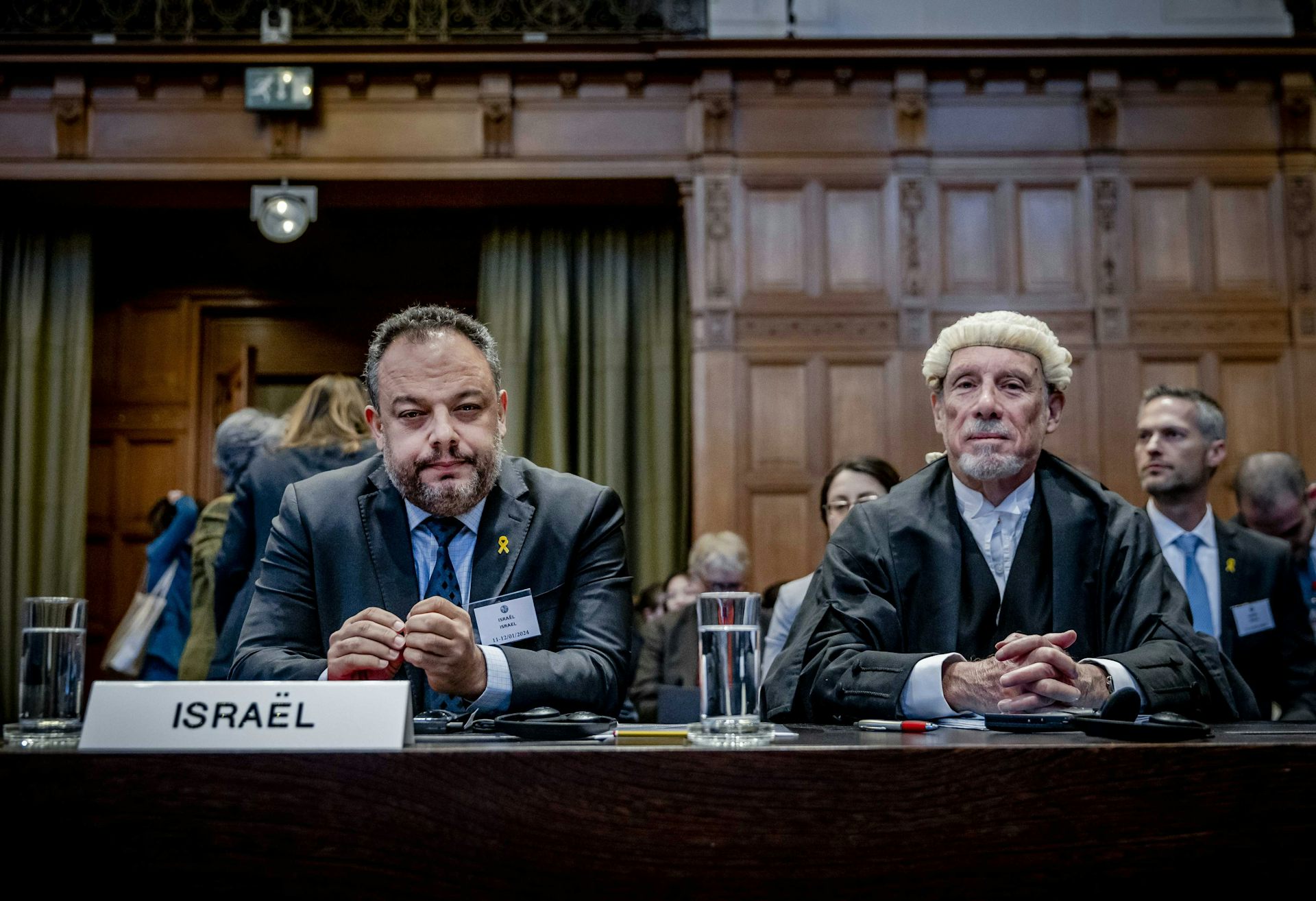 Two men, one wearing a lawyer's wig, sit behind a small plaque reading 'Israel' in a courtroom