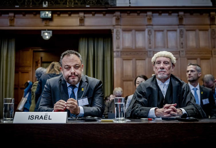 Two men, one wearing a lawyer's wig, sit behind a small plaque reading 'Israel' in a courtroom