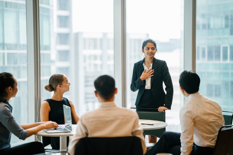 Woman standing in front of room addressing colleagues seated at a table