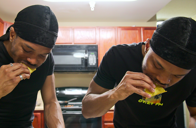 Twin men in identical black clothes stand in a kitchen eating tacos
