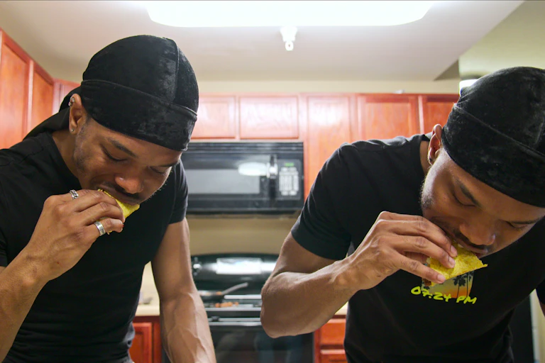 Twin men in identical black clothes stand in a kitchen eating tacos