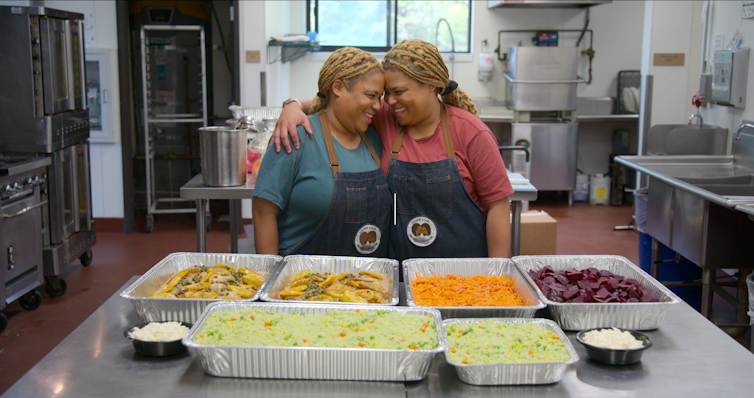 Two women with blonde dreadlocks in a commercial kitchen with various food trays in front of them