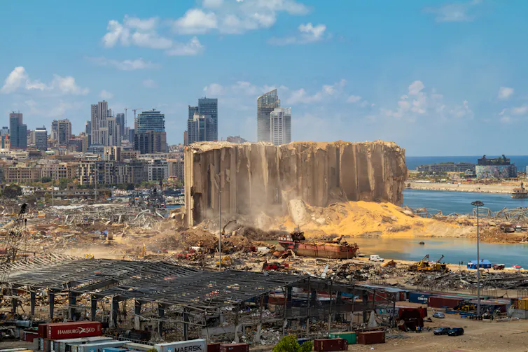 Rubble surrounding the remains of a large building next to the sea with tower blocks in the background.