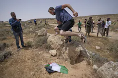A man in shorts and sandals jumps on a Palestinian flag lying on the ground as people surround him.
