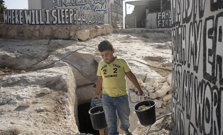 A boy in a yellow T-shirt carries two buckets from a cave.