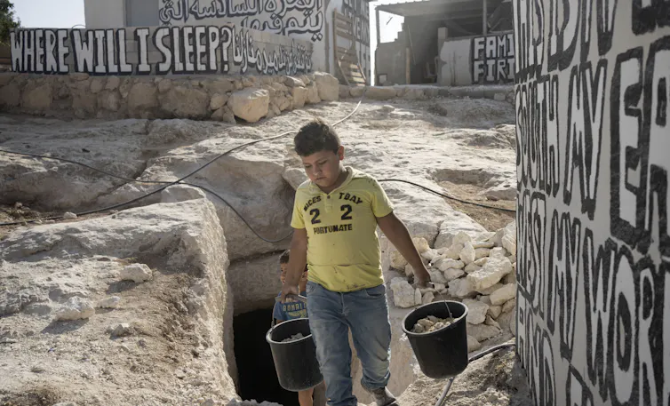 A boy in a yellow T-shirt carries two buckets from a cave.