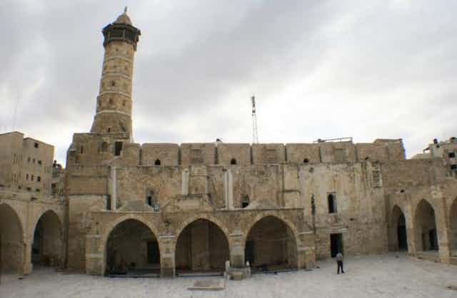 An ancient stone mosque with a large courtyard in front and a tall minaret.