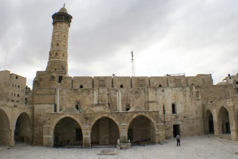 An ancient stone mosque with a large courtyard in front and a tall minaret.