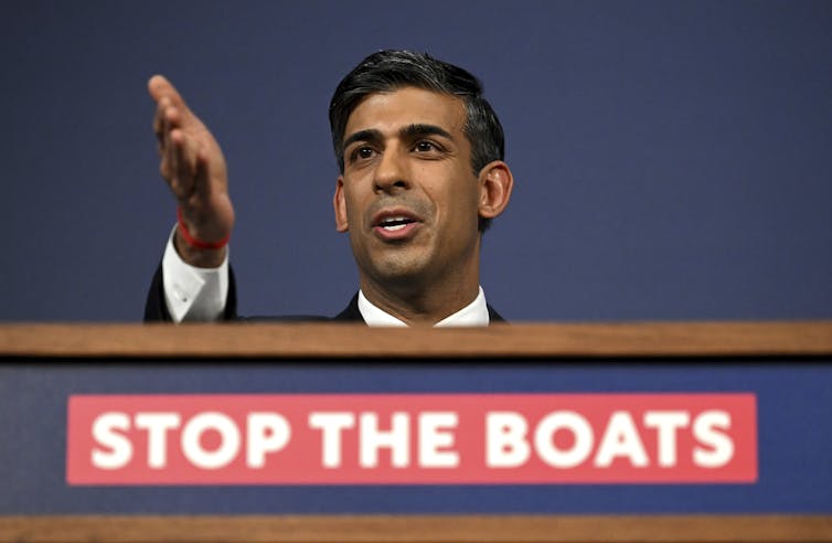 A man gestures behind a podium emblazoned with the words Stop the Boats.