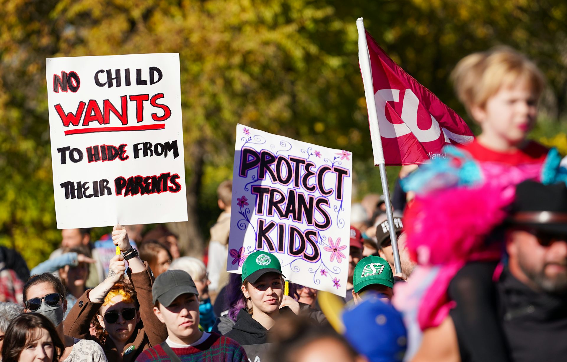 A placard at a rally says 'protect trans kids' next to a CUPE flag.