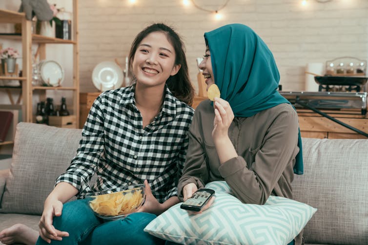 Two young women sitting on a couch eating chips.