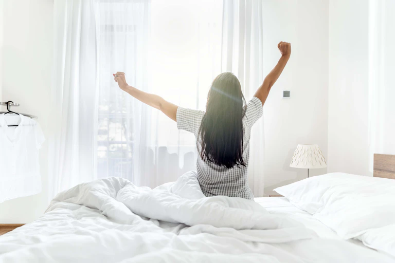 Woman with long black hair stretching after awakening, looking toward a bright window.