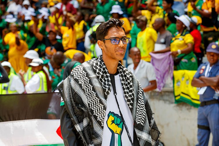A bespectacled young man wearing a keffiyeh and a T-shirt showing support for Palestine.