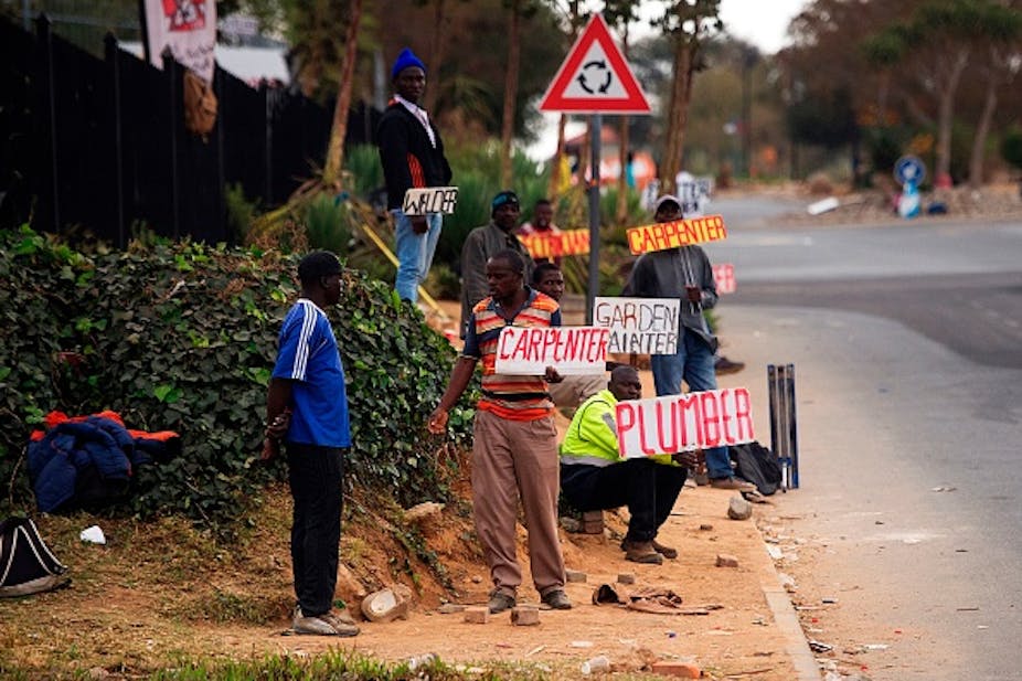 Men stand at a roadside, holding placards. In the background is a traffic sign with arrows making a circle shape.