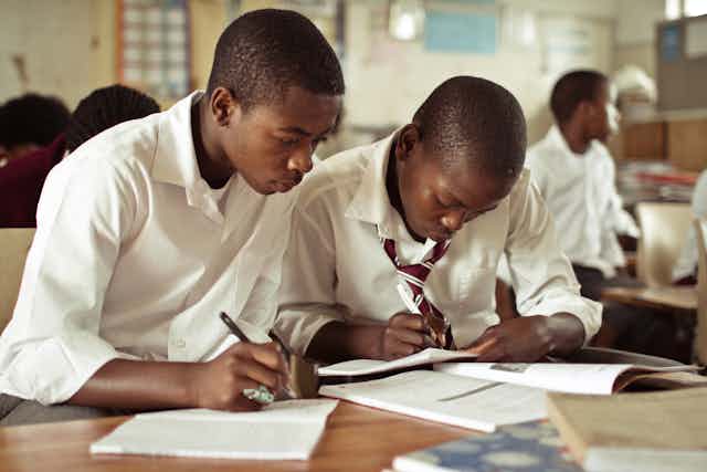 Two boys writing in books while looking at a textbook