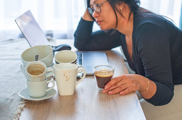 A tired woman looks at her laptop with multiple finished cups of coffee.