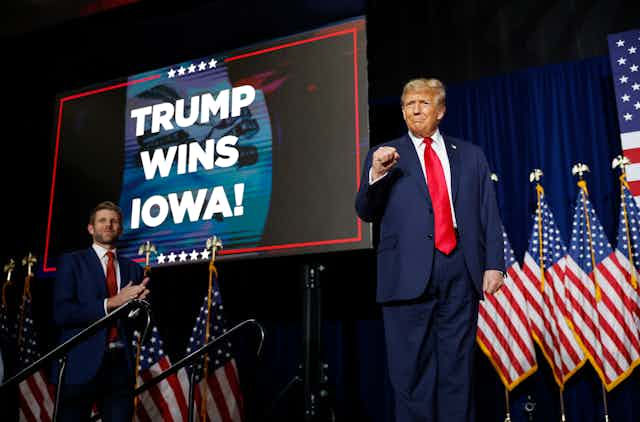 Donald Trump pumps his fist in the air and stands in front of a row of American flags. Behind him a screen says 'Trump wins Iowa!' and his son Eric Trump, also wearing a blue suit and red tie, claps his hands. 