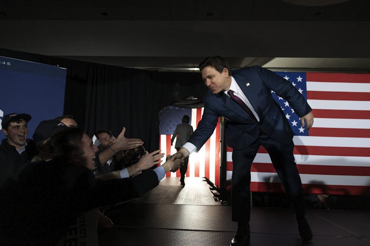 Ron DeSantis wears a navy suit and bends down to shake people's hands as he walks on a stage. He stands in front of two large American flags.