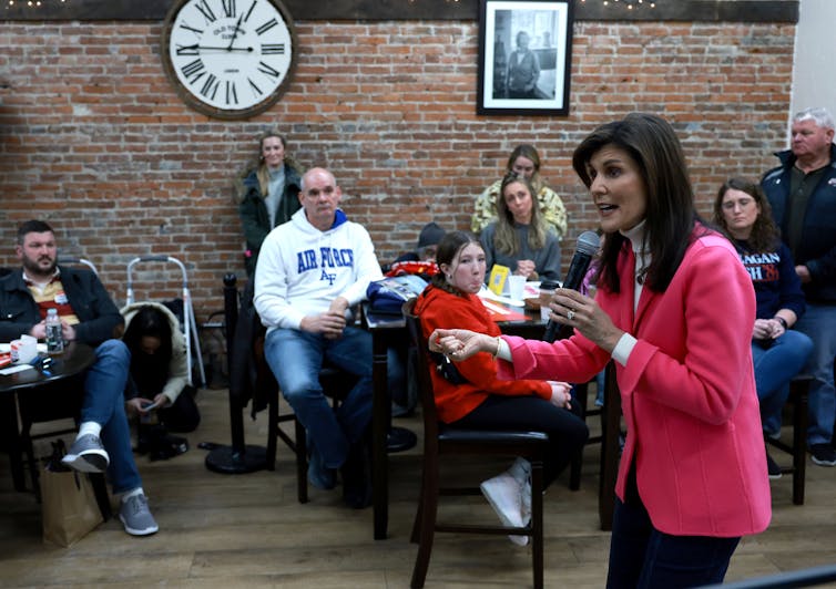 Nikki Haley wears a pink blazer and speaks into a microphone, as she stands in front of a group of people sitting at tables watching her.