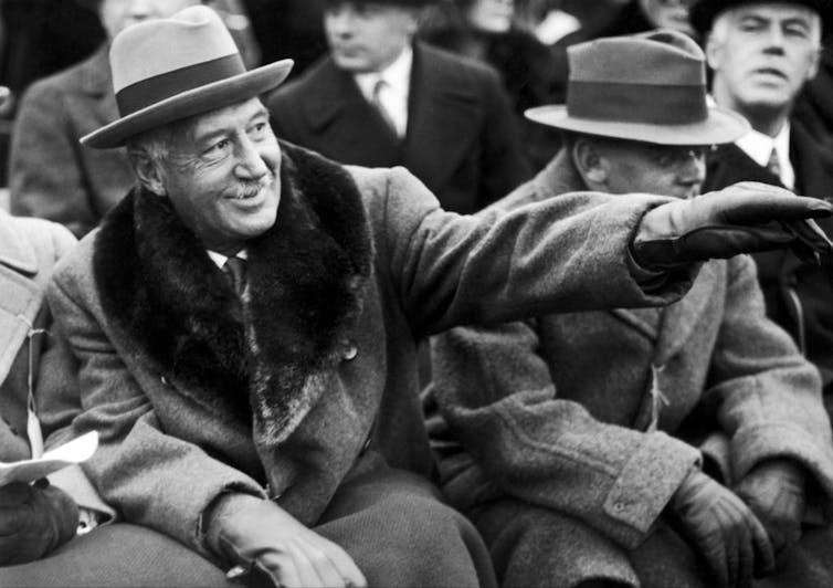 Black and white photo of mustached man wearing heavy winter coat and a fedora.