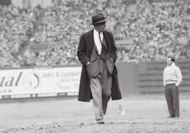 Black and white photo of man wearing fedora and long overcoat patrolling the football field with his hands in his jacket pockets.