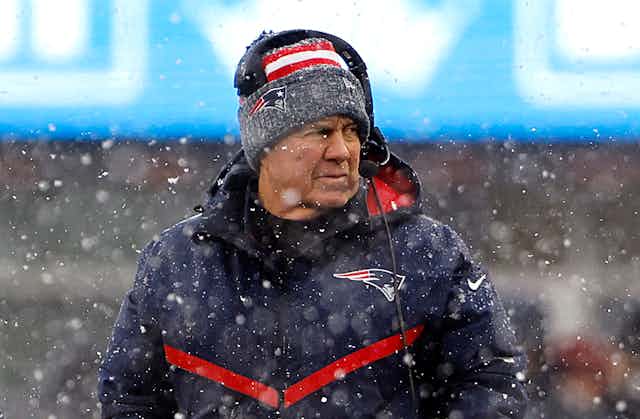 Older man wearing a headset, a knit cap and a winter coat glances to his left surrounded by snow flurries.