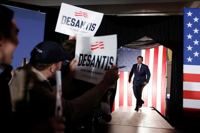 Ron DeSantis walks on a stage as people wave DeSantis signs. Two American flags are also seen near a stage. 