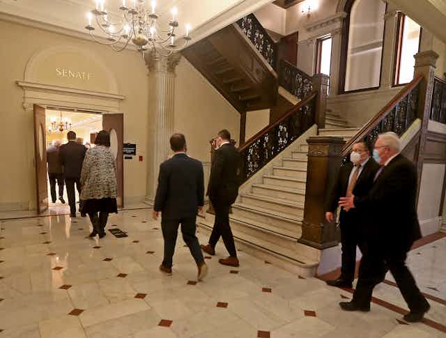people wearing suits walk under chandelier through door marked 'SENATE'