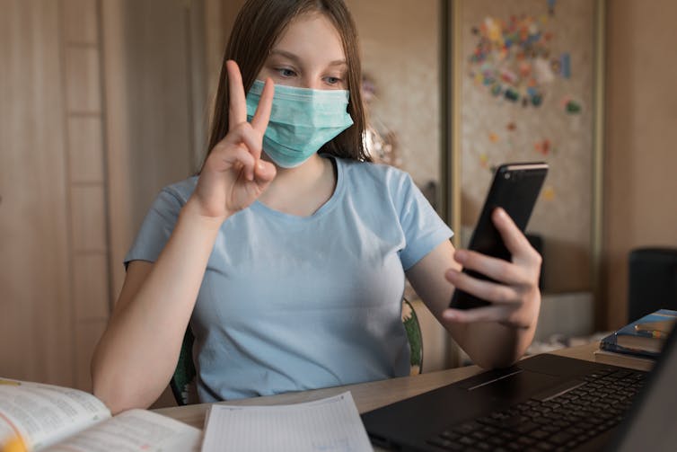 a teen girl wearing a face mask makes a peace sign to her phone