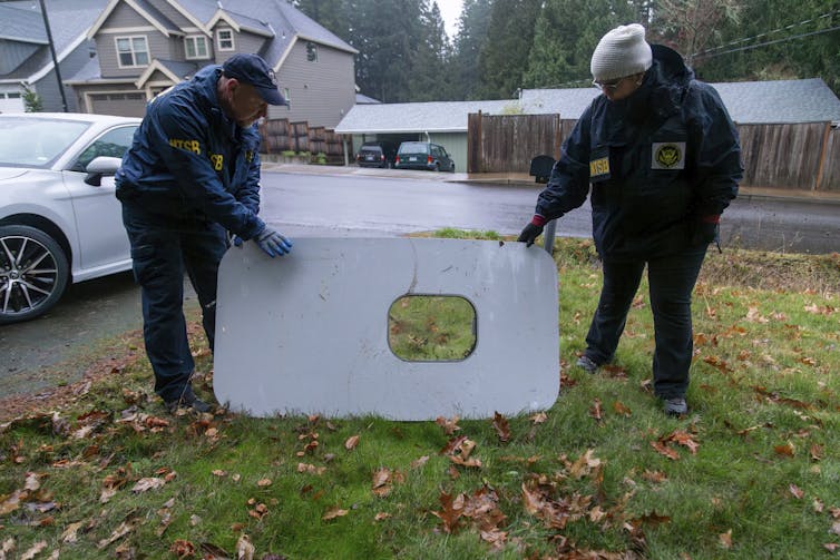 Two agents with the NTSB display a large, door-sized piece of plane detritus on a suburban lawn.