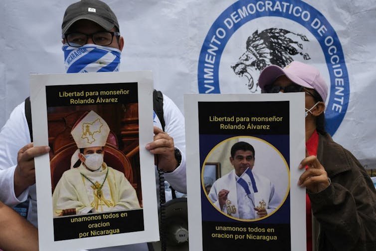 Two people in baseball hats hold posters with pictures of a man in clerical robes.