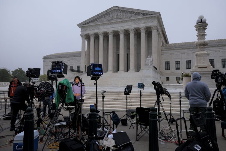 Multiple professional looking video cameras and other equipment are stationed closely together outside the Supreme Court on a grey day. A few people also stand with the equipment, with one woman looking toward a camera.