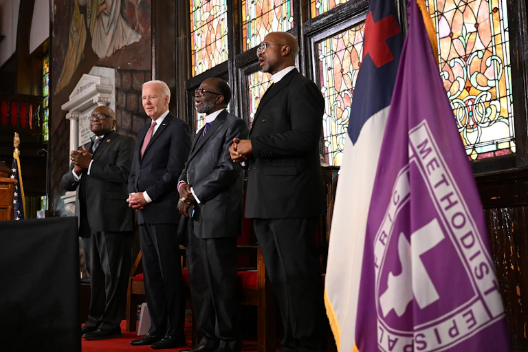 A white man is next to three Black men in front of a brightly colored, stained glass window.