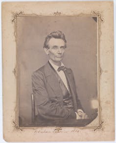 In this black-and-white photograph, a white man dressed in a dark suit sits in a chair with his hands on his lap.