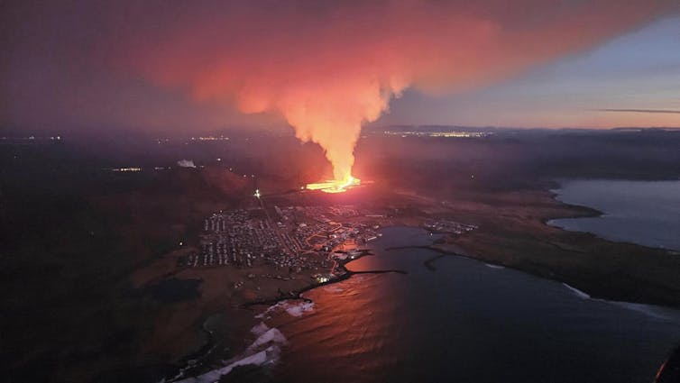 The glow from lava lights up the sky with a town nearby in front of it.