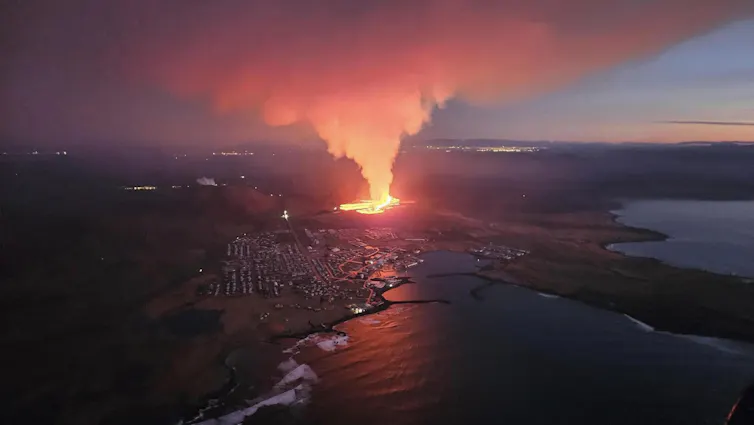The glow from lava lights up the sky with a town nearby in front of it.
