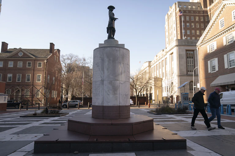 A small man atop a large pedestal in the middle of an urban, paved park.