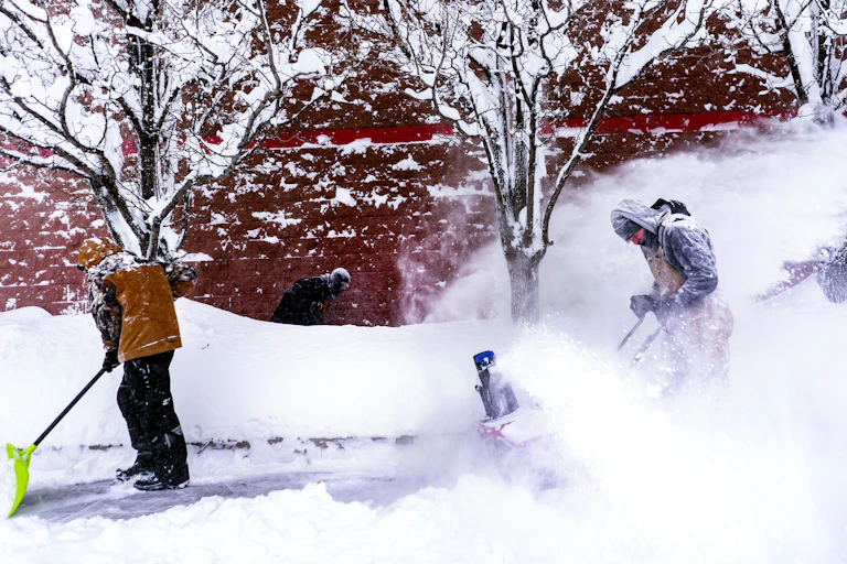 Workers put their heads down against the bitter cold wind as they shovel and blow snow off a sidewalk.