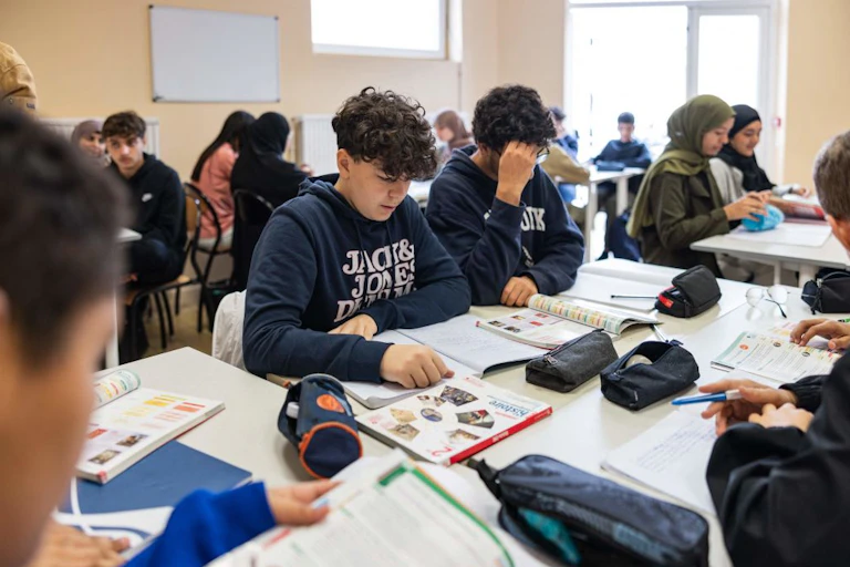 Students seated at white tables look down at their work in a room with beige walls.