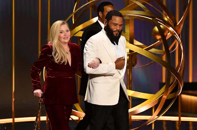 Christina Applegate walks on stage, arm in arm with event host Anthony Anderson. Applegate has long blonde hair and wears a long sleeved velvet evening gown in a dark red shade. She leans on a walking stick. Anderson has a beard and wears a tuxedo with a white jacket.
