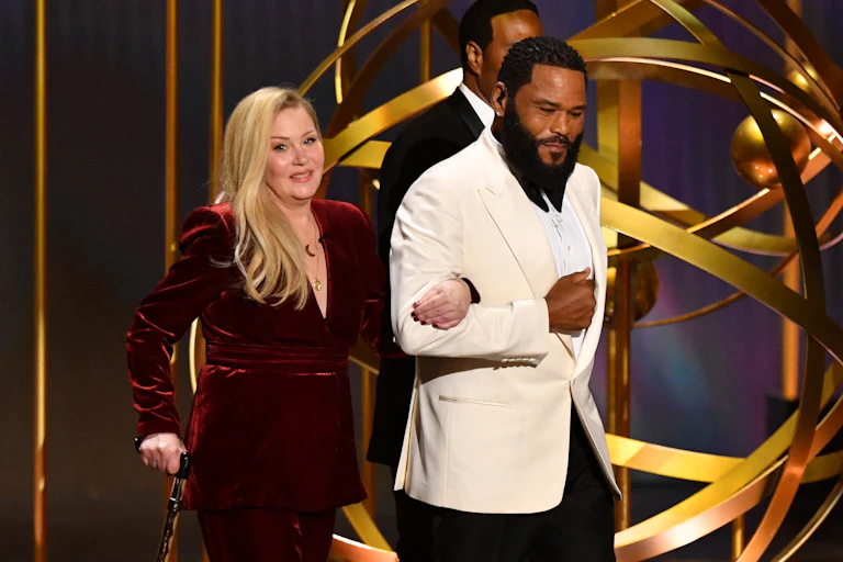 Christina Applegate walks on stage, arm in arm with event host Anthony Anderson. Applegate has long blonde hair and wears a long sleeved velvet evening gown in a dark red shade. She leans on a walking stick. Anderson has a beard and wears a tuxedo with a white jacket.