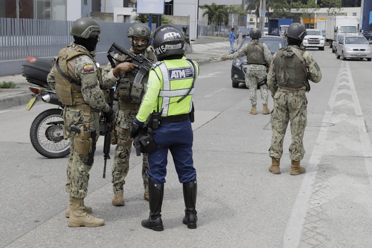 Armed soldiers blocking a road, talking to a motorbike driver.