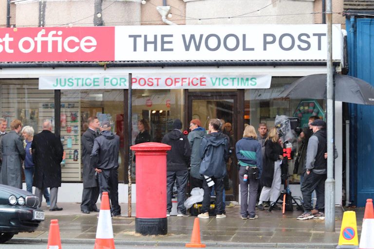 A film crew and actors on a pavement outside a shop.